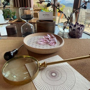 May include: A collection of objects on a table, including a brass magnifying glass, a wooden bowl with a pink crystal, a glass hourglass, and a card with Hebrew text. The scene is illuminated by natural light, suggesting a workspace or display.