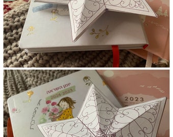 May include: A white paper star with a detailed, intricate design in black ink. The star is folded and appears three-dimensional. The star is sitting on top of a closed book with a floral design.