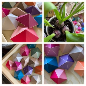 Peut inclure: Un assemblage de pyramides géométriques en papier aux couleurs vives : rose, rouge, bleu, violet et beige. Certaines pyramides sont disposées sur une surface en bois, d'autres dans un plateau en bois. Une plante en pot avec des feuilles vertes et des bulbes est également visible.