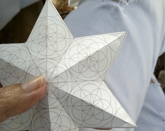 Könnte beinhalten: Ein weißer Papierstern mit einem geometrischen Muster aus überlappenden Kreisen. Der Stern wird von einer Hand gehalten.