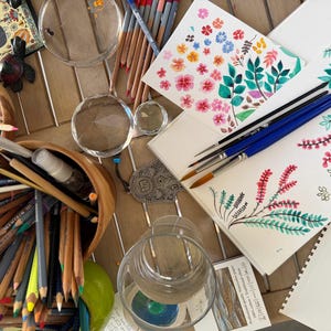 Könnte beinhalten: Draufsicht auf Kunstzubehör auf einem Holztisch. Aquarellmalereien von Blumen und Blättern liegen neben Pinseln mit blauen Griffen. Buntstifte, eine Holzschale und transparente Glasobjekte sind ebenfalls zu sehen.