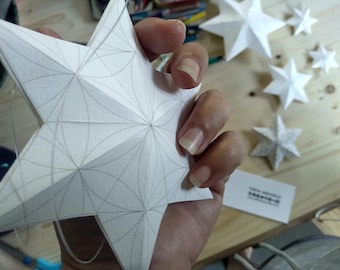 Könnte beinhalten: Ein weißer Papierstern mit geometrischem Muster wird von einer Hand gehalten. Der Stern ist gefaltet und hat eine Schnur daran befestigt. Im Hintergrund befinden sich weitere weiße Papiersterne.