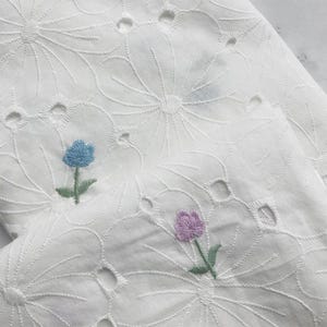 May include: A white fabric featuring an intricate eyelet embroidery pattern of large floral shapes. The textile is accented with small, individual embroidered flowers in light blue and soft purple, each with green stems and leaves.