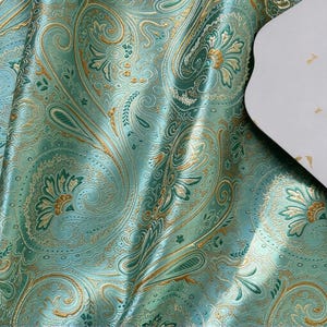 May include: A fabric featuring a repeating paisley pattern in shades of teal, light green, and metallic gold. The surface has a smooth, reflective sheen with intricate floral and teardrop motifs woven throughout the textile design.