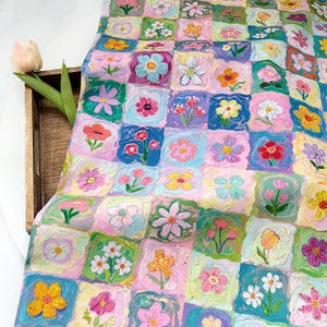 May include: A fabric sheet featuring a repeating grid pattern of hand-painted style flowers in various colors including pink, blue, yellow, and green. Each square contains a unique floral illustration with visible brushstroke textures.