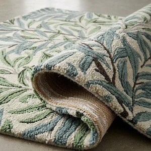 Op de afbeelding: Een getuft karpet met een botanisch patroon van wilgenbladeren in saliegroen, stofblauw en donkerbruin op een gebroken witte achtergrond. Het kleed is gedeeltelijk omgevouwen, waardoor de gestructureerde, geweven jute achterkant zichtbaar is.