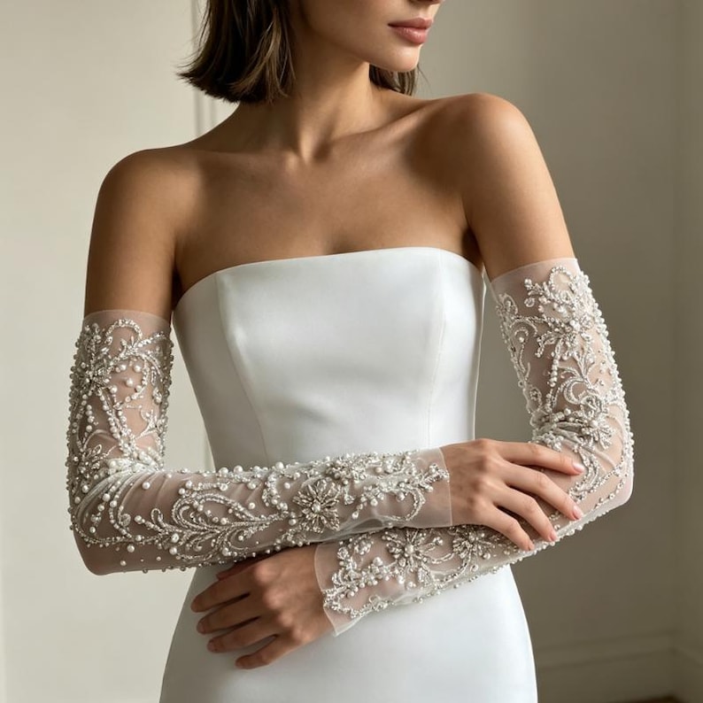 May include: A strapless white gown featuring long, sheer sleeves made of tulle. The sleeves are heavily embellished with intricate silver embroidery, clear crystals, and small white pearls arranged in ornate, swirling floral patterns.