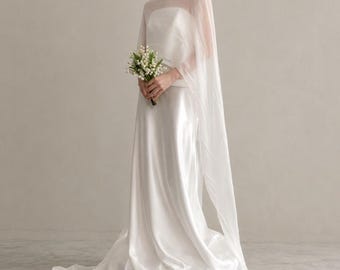 May include: A floor-length white satin gown featuring a sleeveless bodice and a sheer, flowing cape overlay that drapes from the shoulders. The garment is paired with a small bouquet of white lily of the valley flowers held at the waist.