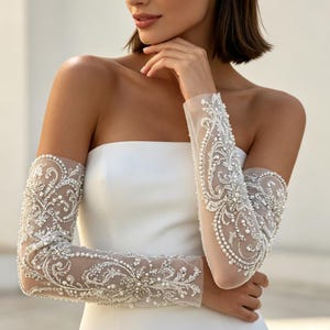 May include: A pair of sheer, elbow-length bridal sleeves featuring intricate white embroidery, silver thread detailing, and scattered white pearls. The sleeves are form-fitting and extend from the upper arm to the wrist, showcasing a dense, ornate floral pattern.