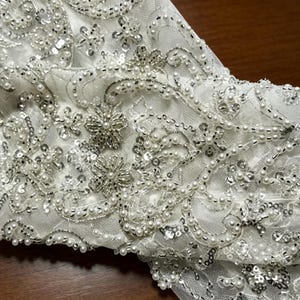 May include: A white sheer fabric overlay featuring intricate embroidery, silver sequins, and small white beads arranged in floral and swirling patterns. The embellishments cover the surface, creating a textured, reflective, and detailed design.
