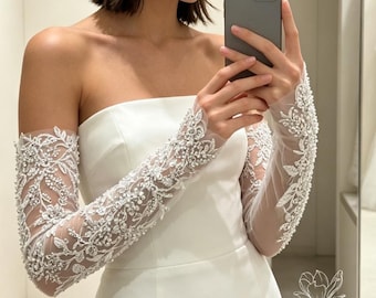 May include: A strapless white gown featuring detachable long sheer sleeves adorned with intricate silver embroidery, clear sequins, and white beads in a floral vine pattern. The sleeves extend from the upper arm to the wrist, contrasting with the smooth fabric.