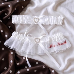 May include: Two white elastic garters featuring a central white satin ribbon bow adorned with a clear rhinestone heart charm. The wider bottom garter includes a ruffled tulle trim and the embroidered red text Mrs. Simith on the right side.