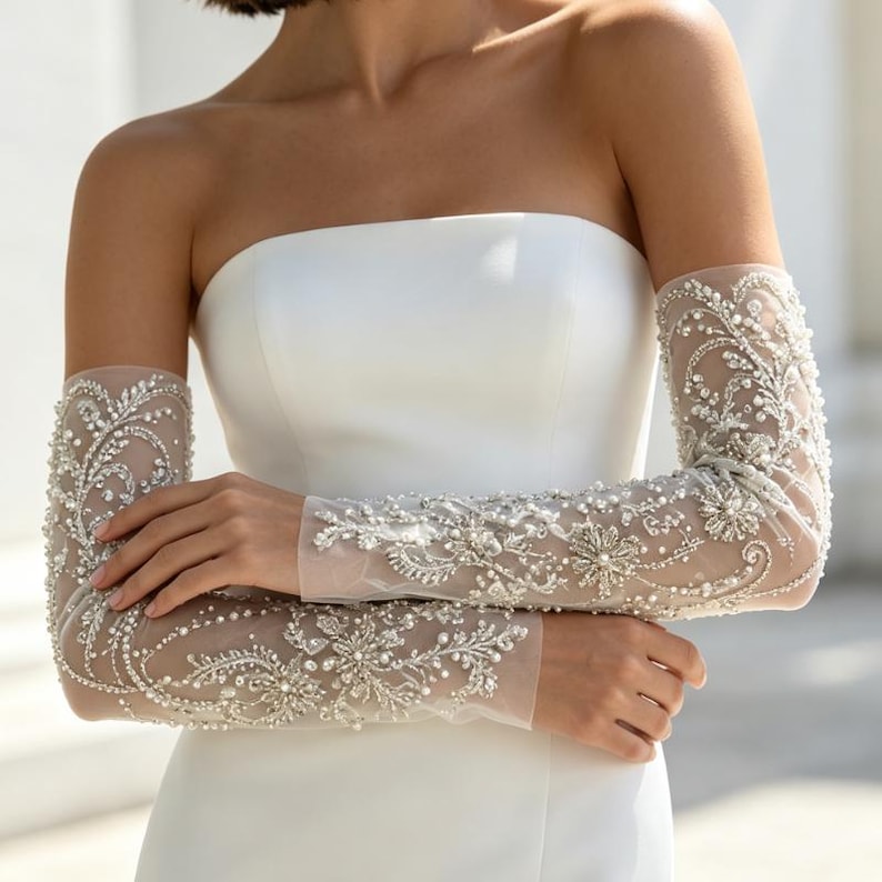 May include: A pair of sheer, elbow-length bridal gloves featuring intricate silver embroidery, clear crystals, and small white pearls in a swirling floral pattern. The gloves are worn with a strapless, solid white gown with a smooth, matte finish.