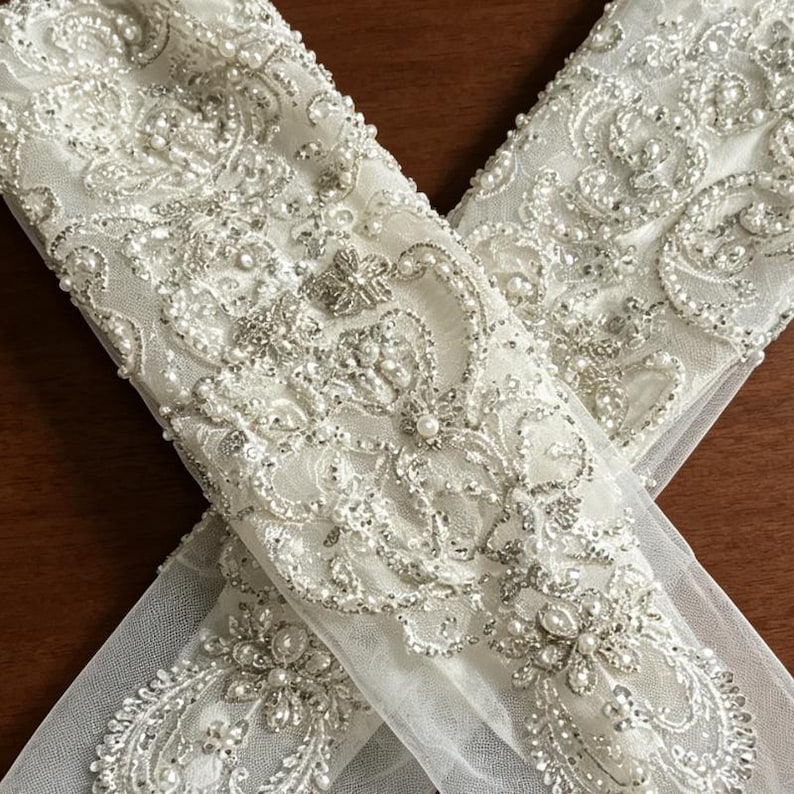 May include: A pair of sheer white tulle gloves featuring intricate silver embroidery, clear sequins, and small white faux pearls arranged in ornate floral and scroll patterns that extend along the length of the fabric in a dense, decorative design.