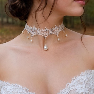 Może przedstawiać: Biały koronkowy naszyjnik typu choker z misternym wzorem kwiatowym i delikatnymi, wykończonymi w ząbki brzegami. Projekt zawiera kilka małych, zwisających białych pereł wzdłuż dolnej krawędzi oraz większą perłę w kształcie łzy pośrodku.