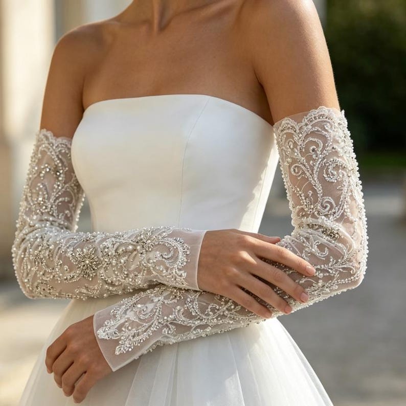 May include: A strapless white gown paired with long, sheer sleeves featuring intricate white lace embroidery, silver beadwork, and small pearls. The sleeves extend from the upper arm to the wrist, showcasing a dense, ornate floral pattern.