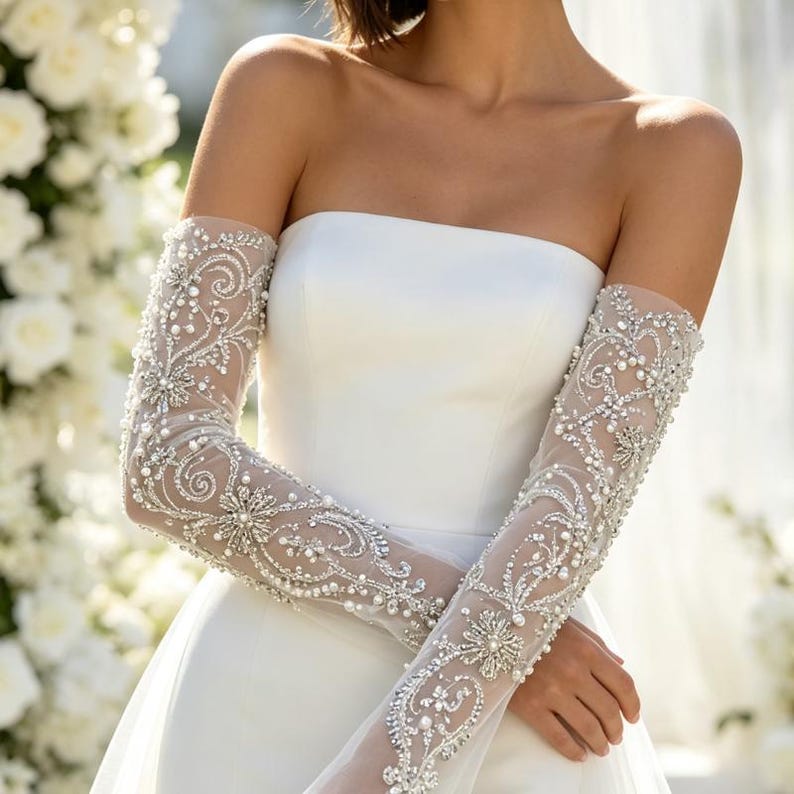 May include: A strapless white gown paired with long, sheer, detachable sleeves. The sleeves are crafted from a translucent mesh fabric and are heavily embellished with intricate silver embroidery, clear crystals, and small white pearls.