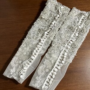 May include: A pair of white sheer lace gloves featuring intricate floral embroidery, silver beadwork, and sequins. Each glove is detailed with a vertical row of small, round, fabric-covered buttons running along the length of the forearm.
