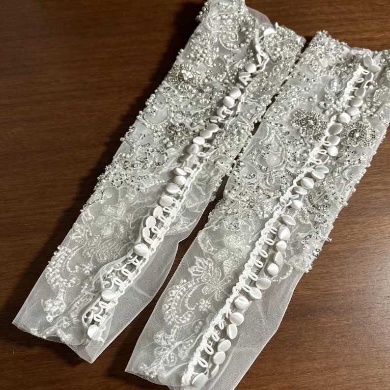 May include: A pair of white sheer lace gloves featuring intricate floral embroidery, silver beadwork, and sequins. Each glove is detailed with a vertical row of small, round, fabric-covered buttons running along the length of the forearm.