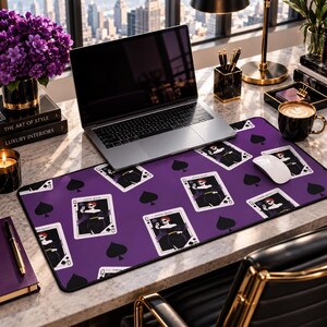 May include: A rectangular desk mat featuring a repeating pattern of playing cards depicting a queen figure in a black dress and sunglasses, interspersed with black spade symbols on a solid purple background with a black stitched border.