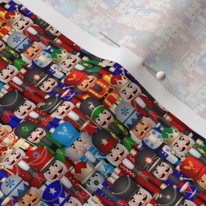 May include: A fabric textile featuring a repeating pattern of nutcracker figures in various uniforms, including red, blue, green, and gold colors. The nutcrackers have painted facial features, mustaches, and hats. A white fabric edge is visible.