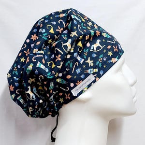 Puede incluir: Un gorro quirúrgico azul marino con un estampado navideño festivo. El gorro presenta varias ilustraciones con temas navideños, como soldados de juguete, caballos balancín y bastones de caramelo. El texto "TheCraftyPharmerSC" es visible en el lateral.