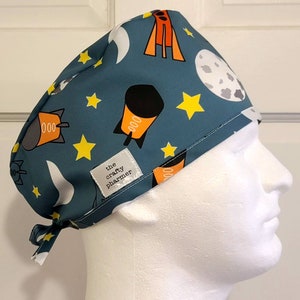 Pode incluir: Uma touca cirúrgica azul-petróleo com um padrão com tema espacial. O design inclui foguetes, estrelas, luas e planetas em laranja, amarelo e branco. A touca tem uma etiqueta que diz "the crafty pharmer".