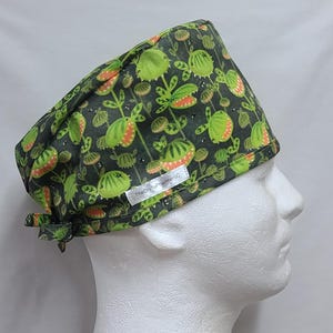 May include: Green scrub hat with a print of cartoon carnivorous plants. The hat has a tie at the back and a label that reads "The Crafty Hamsters SG".