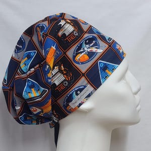 Puede incluir: Un gorro quirúrgico azul marino con un patrón repetido de imágenes temáticas de Star Wars. El diseño incluye cazas estelares X-wing, cazas TIE y otros gráficos espaciales en tonos azules, naranjas y grises. El gorro tiene un cierre de lazo.