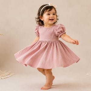 May include: A dusty rose-colored dress with short puff sleeves and a gathered skirt. The dress features a round neckline and a fitted bodice. A small cream-colored bow headband is worn on the head, complementing the simple design of the garment.
