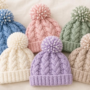 Puede incluir: Una colección de seis gorros tejidos en azul, rosa, verde salvia, crema, lavanda y beige. Cada gorro presenta un patrón de tejido de trenzas grueso, un puño acanalado y un pompón grande y texturizado en la parte superior a juego.