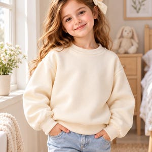 May include: A cream-coloured crewneck sweatshirt with long sleeves and ribbed cuffs, paired with light blue denim jeans. The sweatshirt features a simple, solid-coloured design with a rounded neckline and a relaxed fit, worn with a cream hair bow.
