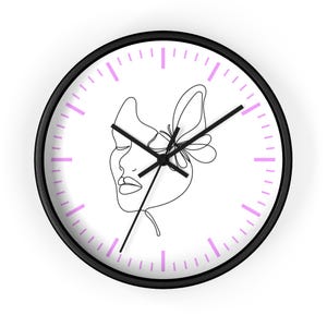 Könnte beinhalten: Eine runde Wanduhr mit schwarzem Rahmen und weißem Zifferblatt. Die Uhr zeigt eine schwarze Strichzeichnung eines menschlichen Gesichts mit einem Schmetterling an der Seite. Das Zifferblatt hat hellviolette Markierungen und schwarze Zeiger.
