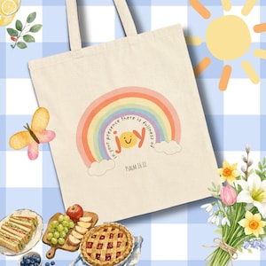 May include: A beige canvas tote bag featuring a colorful rainbow graphic with two clouds at the base. The text reads, "in your presence there is fullness of joy" with a smiley face in the word "joy" and "PSALM 16:11" printed below.
