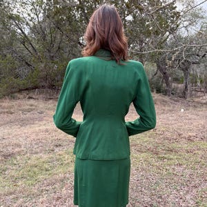 Peut inclure: Un tailleur jupe vert &eacute;meraude vintage. La veste a des manches longues et une coupe ajust&eacute;e, associ&eacute;e &agrave; une jupe assortie jusqu'aux genoux. Le costume est fait d'un tissu lisse et est vu de dos, en ext&eacute;rieur.