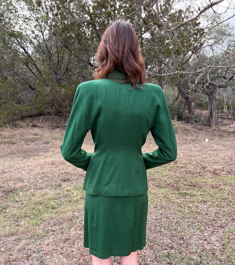 Peut inclure: Un tailleur jupe vert &eacute;meraude vintage. La veste a des manches longues et une coupe ajust&eacute;e, associ&eacute;e &agrave; une jupe assortie jusqu'aux genoux. Le costume est fait d'un tissu lisse et est vu de dos, en ext&eacute;rieur.