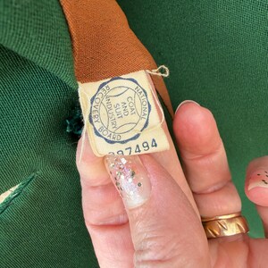Peut inclure: Gros plan sur un v&ecirc;tement vert avec une doublure marron. Une &eacute;tiquette avec le texte "NATIONAL COAT AND SUIT INDUSTRY BOARD" et le num&eacute;ro "297494" est visible. Le v&ecirc;tement est tenu par une personne.