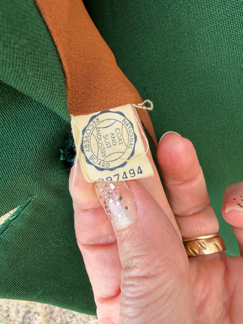Peut inclure: Gros plan sur un v&ecirc;tement vert avec une doublure marron. Une &eacute;tiquette avec le texte "NATIONAL COAT AND SUIT INDUSTRY BOARD" et le num&eacute;ro "297494" est visible. Le v&ecirc;tement est tenu par une personne.
