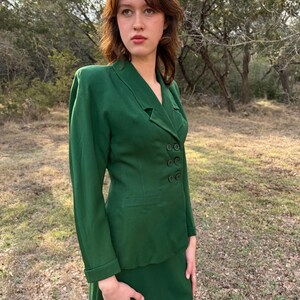 Peut inclure: Un costume deux pi&egrave;ces vert compos&eacute; d'une veste &agrave; double boutonnage et d'une jupe assortie. Le costume est d'un vert vif uni. La veste a des manches longues et une coupe ajust&eacute;e. La jupe arrive au niveau du genou.