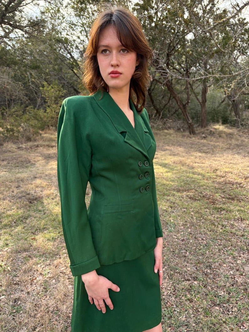 Peut inclure: Un costume deux pi&egrave;ces vert compos&eacute; d'une veste &agrave; double boutonnage et d'une jupe assortie. Le costume est d'un vert vif uni. La veste a des manches longues et une coupe ajust&eacute;e. La jupe arrive au niveau du genou.