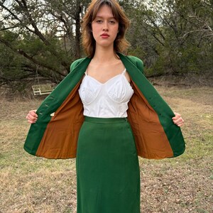Peut inclure: Un tailleur jupe vert vintage avec un blazer assorti. Le blazer a une doublure marron et est ouvert, r&eacute;v&eacute;lant un haut blanc. La tenue est port&eacute;e &agrave; l'ext&eacute;rieur, avec un arri&egrave;re-plan naturel.