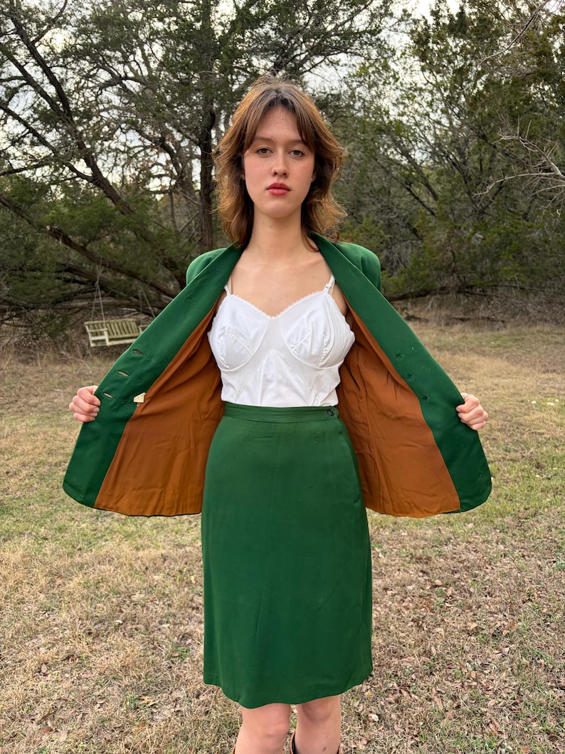 Peut inclure: Un tailleur jupe vert vintage avec un blazer assorti. Le blazer a une doublure marron et est ouvert, r&eacute;v&eacute;lant un haut blanc. La tenue est port&eacute;e &agrave; l'ext&eacute;rieur, avec un arri&egrave;re-plan naturel.
