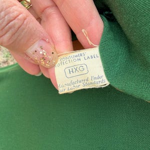 Peut inclure: Gros plan sur un tissu vert avec une &eacute;tiquette attach&eacute;e. L'&eacute;tiquette indique "CONSUMERS PROTECTION LABEL" et "Manufactured Under Fair Labor Standards". L'&eacute;tiquette porte &eacute;galement les lettres "HXG".