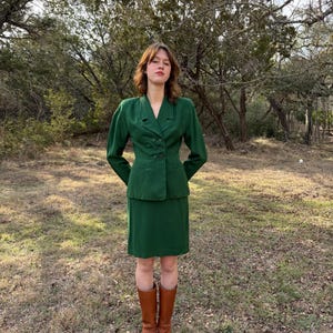 Peut inclure: Un tailleur jupe vert compos&eacute; d'un blazer &agrave; double boutonnage et d'une jupe au genou assortie. L'ensemble est compl&eacute;t&eacute; par des bottes hautes en cuir marron. La tenue est mise en valeur sur un fond d'arbres et d'herbe.