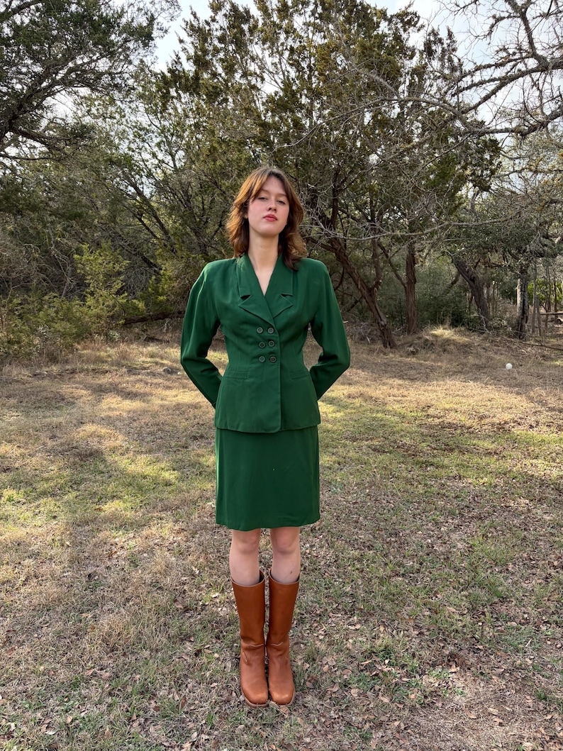 Peut inclure: Un tailleur jupe vert compos&eacute; d'un blazer &agrave; double boutonnage et d'une jupe au genou assortie. L'ensemble est compl&eacute;t&eacute; par des bottes hautes en cuir marron. La tenue est mise en valeur sur un fond d'arbres et d'herbe.