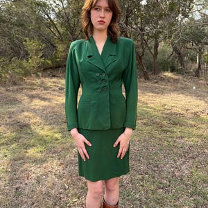 Peut inclure: Un tailleur jupe vert &eacute;meraude vintage. La veste a un design crois&eacute; avec quatre boutons. La jupe arrive au genou. La personne porte des bottes hautes marron. L'arri&egrave;re-plan est un cadre ext&eacute;rieur naturel avec des arbres.