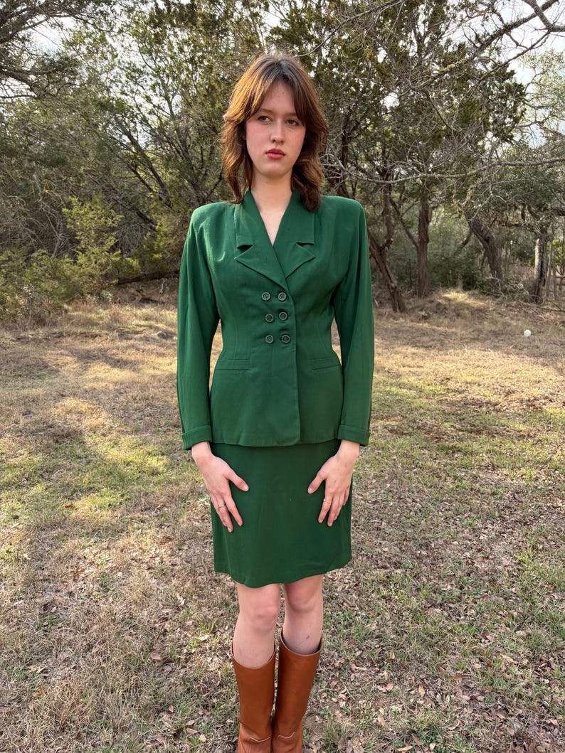 Peut inclure: Un tailleur jupe vert &eacute;meraude vintage. La veste a un design crois&eacute; avec quatre boutons. La jupe arrive au genou. La personne porte des bottes hautes marron. L'arri&egrave;re-plan est un cadre ext&eacute;rieur naturel avec des arbres.