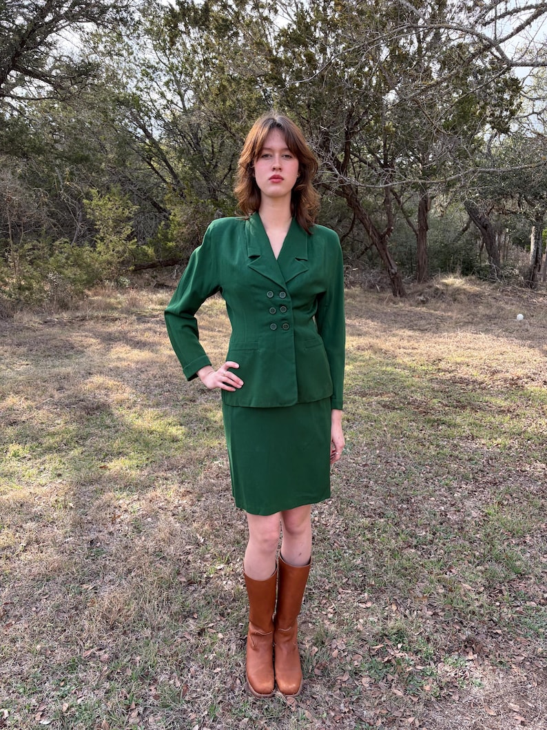 Peut inclure: Un tailleur jupe vert &eacute;meraude vintage, compos&eacute; d'un blazer &agrave; double boutonnage et d'une jupe assortie arrivant au genou. L'ensemble est compl&eacute;t&eacute; par des bottes hautes en cuir marron. La tenue est port&eacute;e en ext&eacute;rieur.