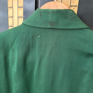 Peut inclure: Gros plan du dos d'un blazer vert fonc&eacute;. Le blazer est suspendu &agrave; un cintre en bois. Le col pr&eacute;sente des taches sombres et de petits trous. Le tissu est une mati&egrave;re unie et textur&eacute;e.