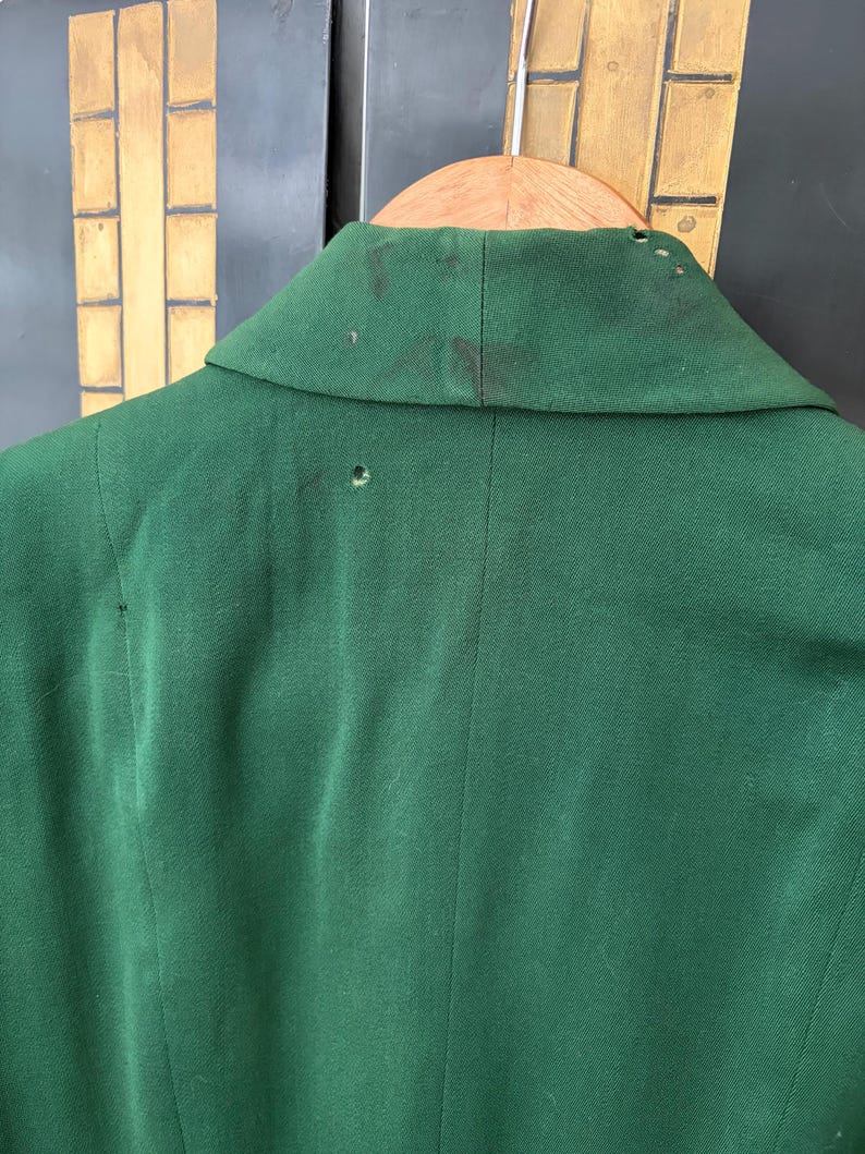Peut inclure: Gros plan du dos d'un blazer vert fonc&eacute;. Le blazer est suspendu &agrave; un cintre en bois. Le col pr&eacute;sente des taches sombres et de petits trous. Le tissu est une mati&egrave;re unie et textur&eacute;e.
